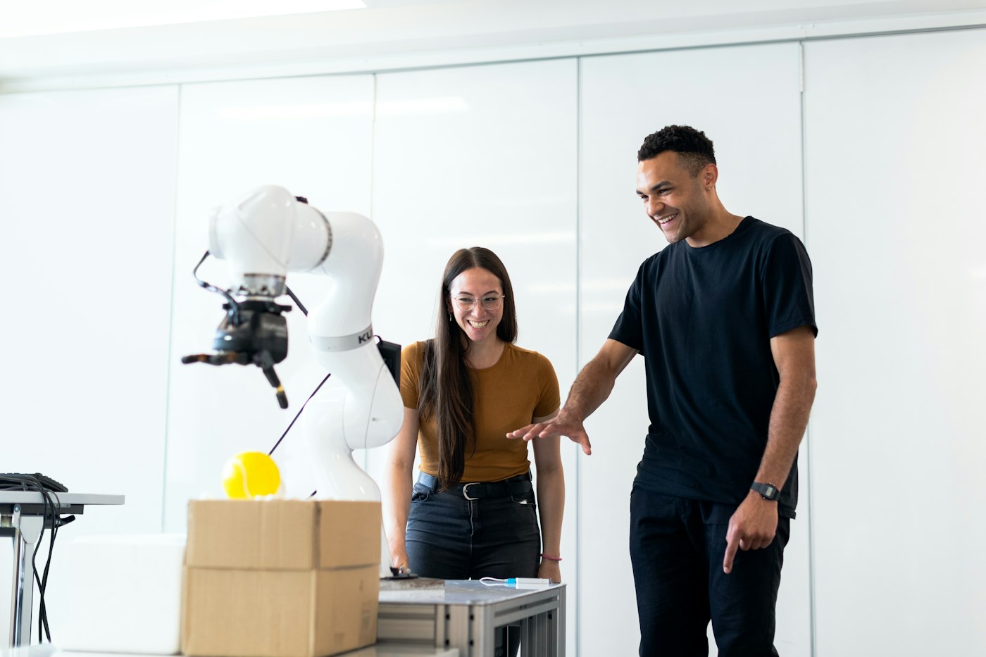 Two people packing orders, smiling, beside a robot arm